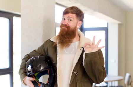 Foto de Hombre de pelo rojo sintiéndose feliz, mostrando aprobación con gesto bien con un casco de moto - Imagen libre de derechos