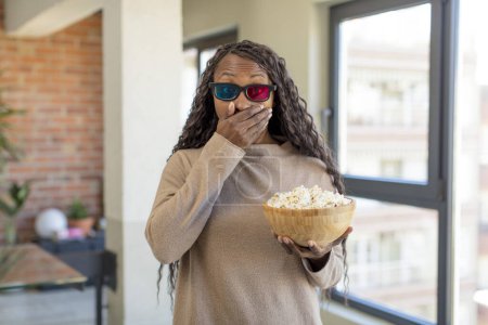 Téléchargez les photos : Noir afro femme couvrant bouche avec une main et choqué ou surpris expression avec popcorns - en image libre de droit