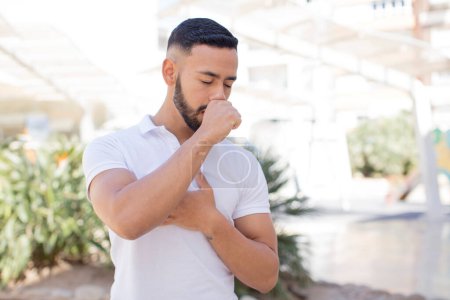 Téléchargez les photos : Jeune homme beau se sentant malade avec un mal de gorge et des symptômes de grippe, toux avec la bouche couverte - en image libre de droit