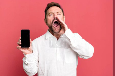 Photo for Middle age man feeling happy,giving a big shout out with hands next to mouth. showing a smartphone - Royalty Free Image