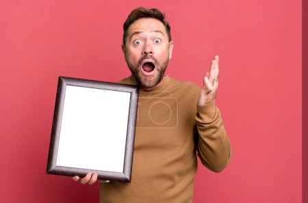 Photo for Middle age man amazed, shocked and astonished with an unbelievable surprise with an empty frame - Royalty Free Image