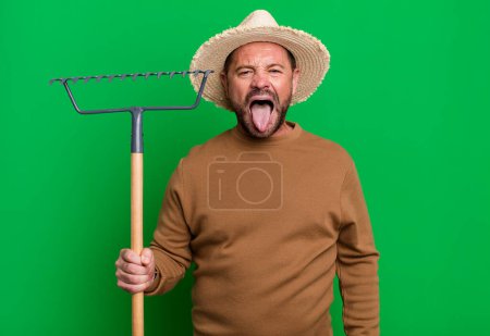 Photo for Middle age man feeling disgusted and irritated and tongue out. farmer with a rake - Royalty Free Image