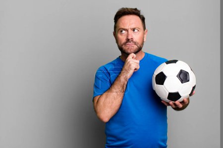 Foto de Hombre de mediana edad pensando, sintiéndose dudoso y confundido. con una pelota de fútbol. concepto de fitness - Imagen libre de derechos