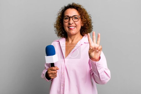 Foto de Bastante mujer de mediana edad sonriendo y buscando amigable, mostrando el número tres. presentador de televisión con un concepto de micrófono - Imagen libre de derechos