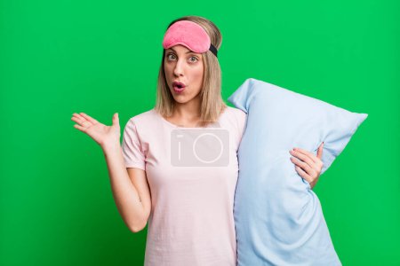 Foto de Mujer rubia bonita mirando sorprendida y conmocionada, con la mandíbula caída sosteniendo un objeto. concepto de pijamas y ropa de dormir - Imagen libre de derechos