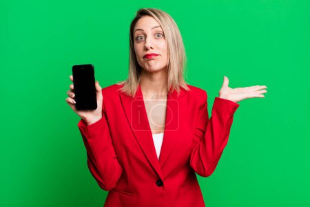 Foto de Bonita mujer rubia sintiéndose desconcertada y confundida y dudando. empresaria mostrando una pantalla de teléfono - Imagen libre de derechos