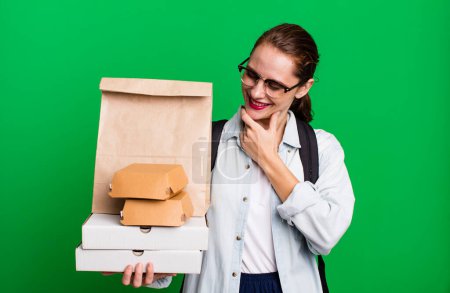 Foto de Mujer hispana bonita sonriendo con una expresión feliz y segura con la mano en la barbilla. entrega comida rápida take away concepto - Imagen libre de derechos