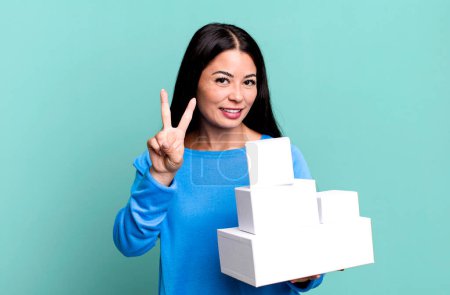 Photo for Hispanic pretty woman smiling and looking friendly, showing number two. with white boxes packagings - Royalty Free Image