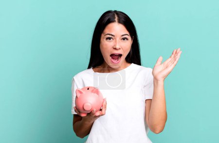 Photo for Hispanic pretty woman feeling happy and astonished at something unbelievable with a piggy bank - Royalty Free Image