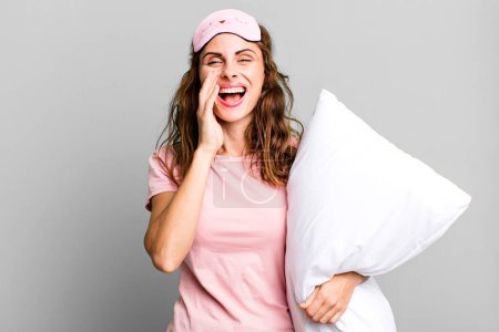 Photo for Hispanic pretty woman feeling happy,giving a big shout out with hands next to mouth wearing pajamas and a pillow - Royalty Free Image