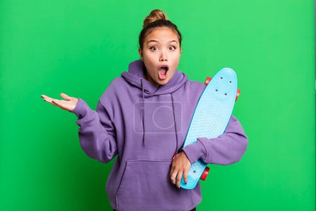 Foto de Hispanic pretty woman looking surprised and shocked, with jaw dropped holding an object. skate boarding concept - Imagen libre de derechos