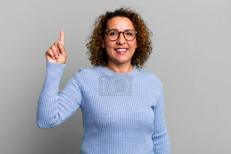 Foto de Mujer hispana de mediana edad sonriendo alegre y felizmente, apuntando hacia arriba con una mano para copiar espacio - Imagen libre de derechos
