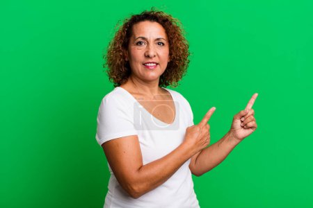 Foto de Mujer hispana de mediana edad sonriendo felizmente y señalando hacia un lado y hacia arriba con ambas manos mostrando el objeto en el espacio de copia - Imagen libre de derechos