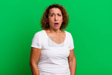 Téléchargez les photos : Femme hispanique d'âge moyen se sentant terrifié et choqué, avec la bouche grande ouverte dans la surprise - en image libre de droit