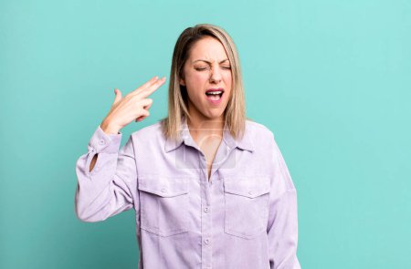 Téléchargez les photos : Blonde femme adulte regardant malheureux et stressé, geste suicidaire faisant signe d'arme à feu avec la main, pointant vers la tête - en image libre de droit