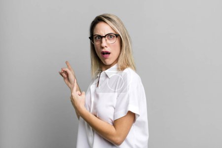 Photo for Blonde adult woman feeling shocked and surprised, pointing to copy space on the side with amazed, open-mouthed look - Royalty Free Image