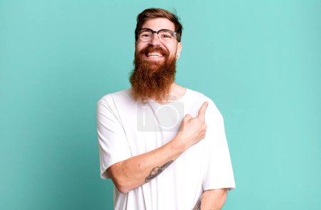 Foto de Barba larga y cabello rojo hombre sonriendo alegremente, sintiéndose feliz y señalando hacia un lado - Imagen libre de derechos