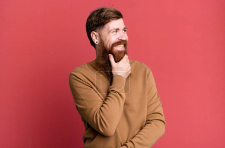 Téléchargez les photos : Barbe longue et cheveux roux homme souriant avec une expression heureuse et confiante avec la main sur le menton - en image libre de droit