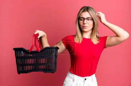 Photo for Young pretty woman smiling happily and daydreaming or doubting. empty shopping basket concept - Royalty Free Image