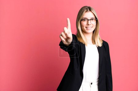 Foto de Joven bonita mujer sonriendo y buscando amigable, mostrando el número uno. concepto de negocio - Imagen libre de derechos
