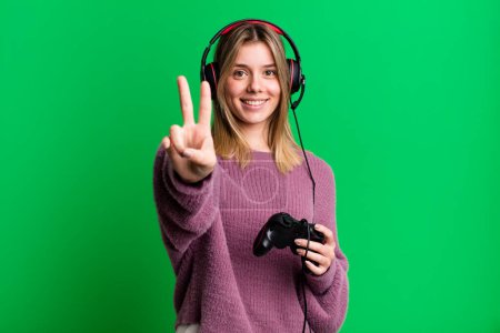 Foto de Joven bonita mujer sonriendo y buscando amigable, mostrando el número dos. gamer con auriculares y controlador - Imagen libre de derechos