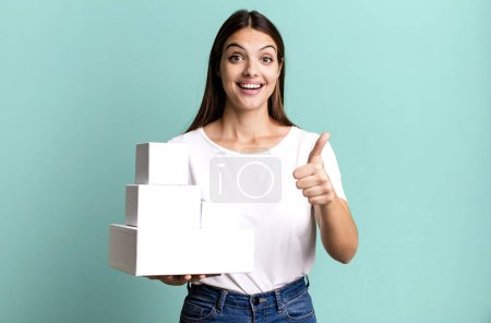 Foto de Joven bonita mujer sintiéndose orgullosa, sonriendo positivamente con los pulgares hacia arriba. cajas blancas en blanco - Imagen libre de derechos