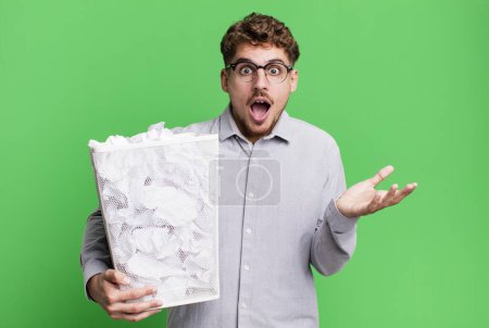 Téléchargez les photos : Jeune homme caucasien adulte étonné, choqué et étonné avec une surprise incroyable avec un concept de poubelle en papier - en image libre de droit