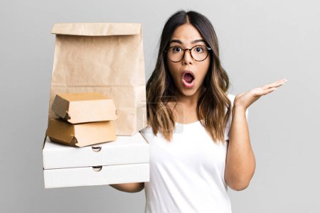Photo for Hispanic pretty woman looking surprised and shocked, with jaw dropped holding an object. delivery and take away fast food concept - Royalty Free Image