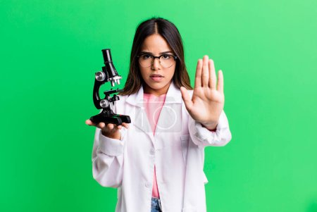 Photo for Hispanic pretty woman looking serious showing open palm making stop gesture. scients student with a microscope - Royalty Free Image