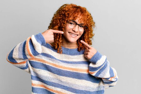 Photo for Redhair pretty woman smiling confidently pointing to own broad smile, positive, relaxed, satisfied attitude - Royalty Free Image