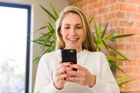 Téléchargez les photos : Jeune adulte jolie femme blonde en utilisant son smartphone à la maison - en image libre de droit