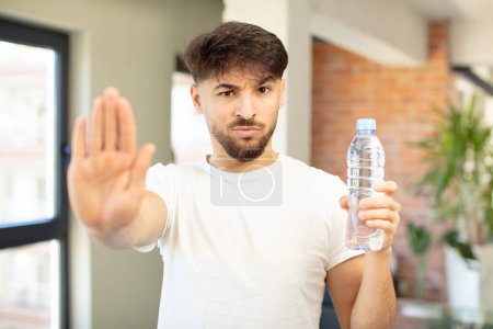 Téléchargez les photos : Jeune homme beau regard sérieux montrant la paume ouverte faisant geste d'arrêt. concept de bouteille d'eau - en image libre de droit