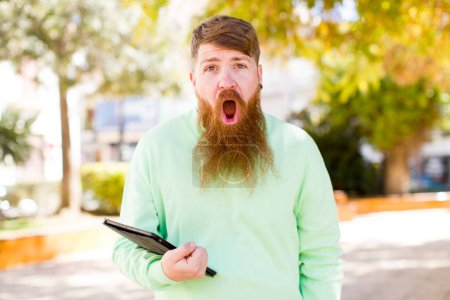 Photo for Red hair young adult man feeling extremely shocked and surprised with a touch screen pad - Royalty Free Image