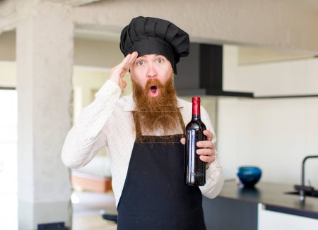 Téléchargez les photos : Homme aux cheveux roux regardant heureux, étonné et surpris avec une bouteille de vin. concept de chef - en image libre de droit
