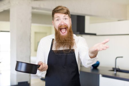 Téléchargez les photos : Homme aux cheveux roux souriant joyeusement et offrant ou montrant un concept dans une cuisine. concept de chef - en image libre de droit