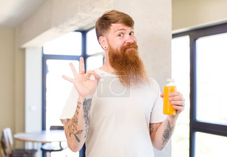 Téléchargez les photos : Homme aux cheveux roux se sentant heureux, montrant son approbation avec un geste correct avec une bouteille de jus d'orange - en image libre de droit