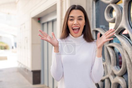 Foto de Mujer adulta bastante joven mirando feliz y emocionado, sorprendido con una sorpresa inesperada con ambas manos abiertas al lado de la cara - Imagen libre de derechos