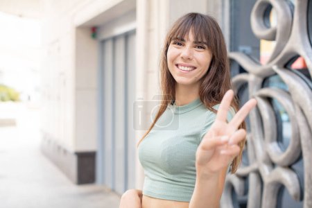 Photo for Pretty young woman smiling and looking happy, carefree and positive, gesturing victory or peace with one hand - Royalty Free Image