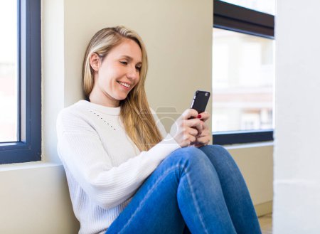 Téléchargez les photos : Jeune adulte jolie femme blonde en utilisant son smartphone à la maison - en image libre de droit