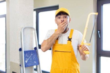 Photo for Young handsome man covering mouth with a hand and shocked or surprised expression. handyman with a saw - Royalty Free Image