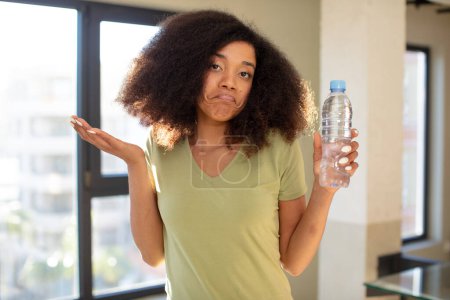 Téléchargez les photos : Jolie afro femme noire haussant les épaules, se sentant confus et incertain. concept de bouteille d'eau - en image libre de droit