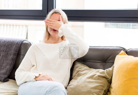 Photo for Young pretty woman covering eyes with one hand feeling scared or anxious, wondering or blindly waiting for a surprise sitting on a couch - Royalty Free Image