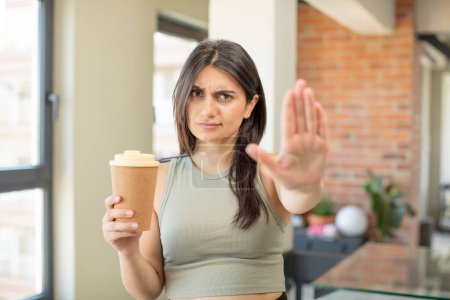 Foto de Mujer joven mirando serio mostrando la palma abierta haciendo gesto de parada. llevar el café - Imagen libre de derechos