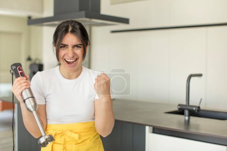 Foto de Mujer joven mirando enojado, molesto y frustrado. chef con licuadora de mano - Imagen libre de derechos