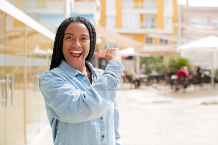 Téléchargez les photos : Afro jolie femme noire se sentant heureuse, satisfaite et puissante, flexion ajustement et biceps musculaires, regardant fort après la salle de gym - en image libre de droit