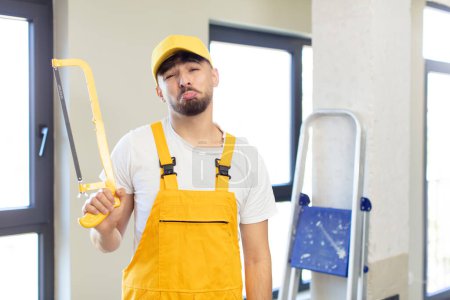 Photo for Young handsome man feeling sad and whiney with an unhappy look and crying. handyman with a saw - Royalty Free Image
