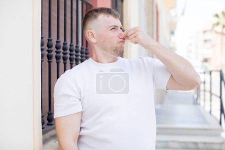 Photo for Handsome man feeling disgusted, holding nose to avoid smelling a foul and unpleasant stench - Royalty Free Image