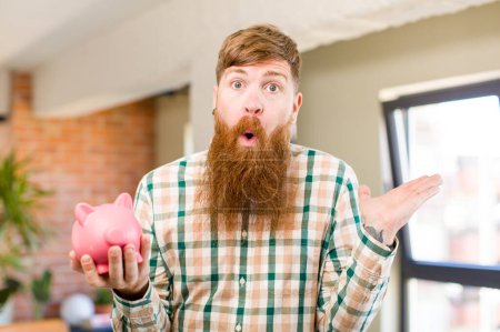 Photo for Red hair man shrugging, feeling confused and uncertain with a piggy bank - Royalty Free Image