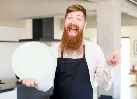 Foto de Hombre de pelo rojo sonriendo alegremente, sintiéndose feliz y señalando hacia un lado con un plato vacío. concepto de chef - Imagen libre de derechos