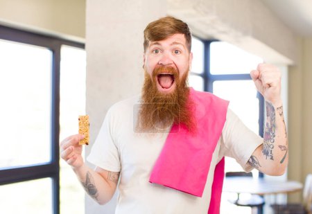 Téléchargez les photos : Homme aux cheveux roux se sentir choqué, rire et célébrer le succès avec une barre de céréales. concept de fitness - en image libre de droit